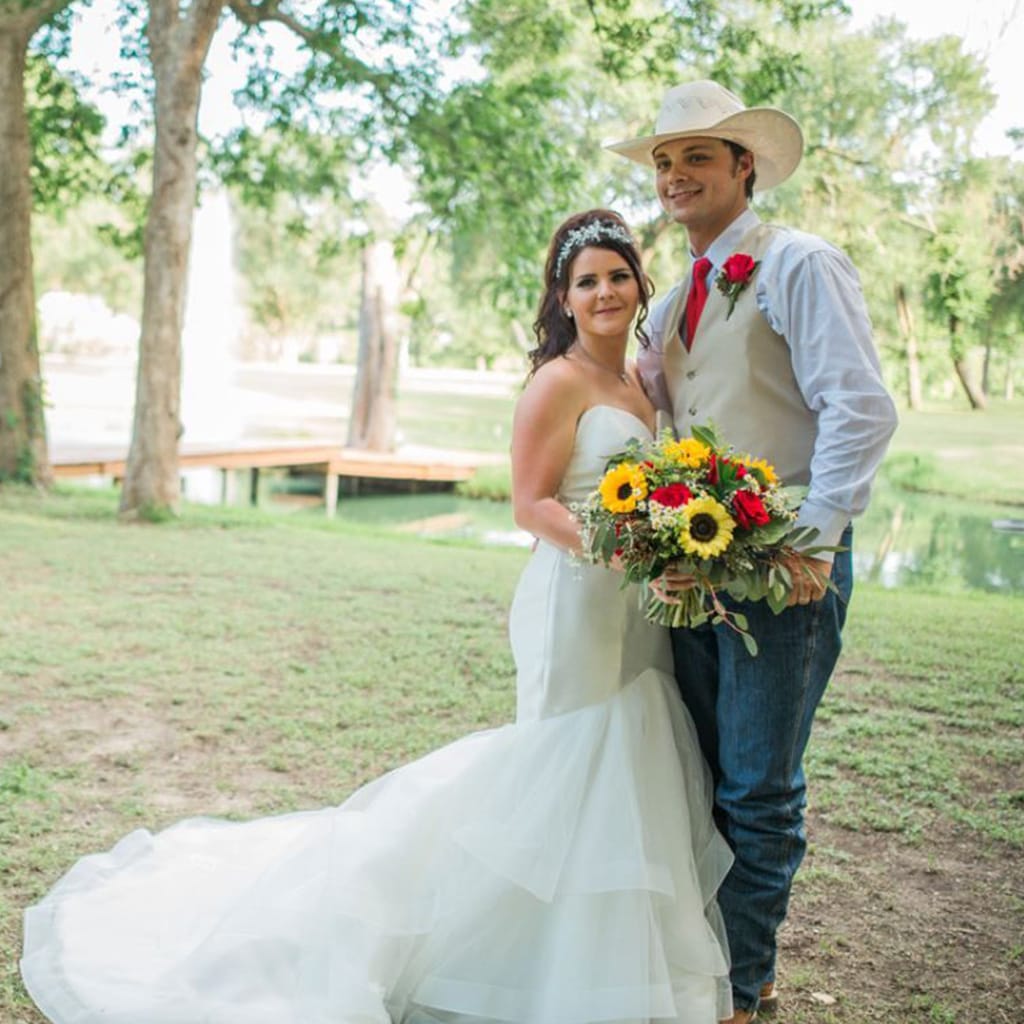 Bride holding a sunflower bouquet beside her groom in a cowboy hat, by a pond