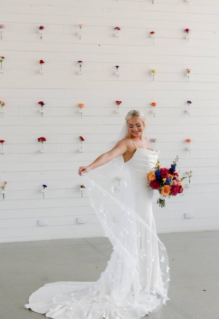 Bride in a white gown holding a bright bouquet in front of a white wall dotted with single flowers