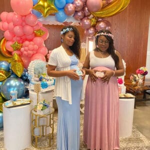 Two expectant mothers in blue and pink dresses and flower crowns in front of a balloon arch