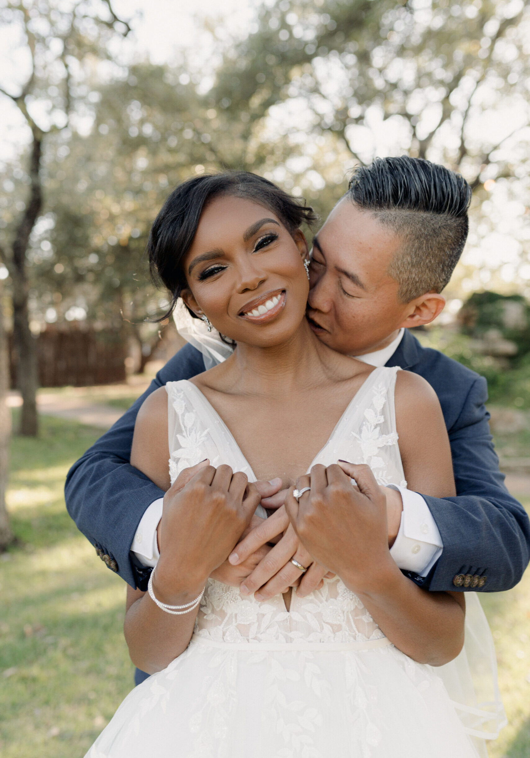 Adaeze & BJ Groom embracing his smiling bride from behind under oak trees