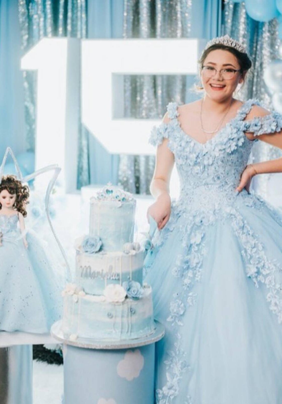 pink-1-1 Young woman in a blue quinceañera gown and tiara beside a tiered cake and a large number 15