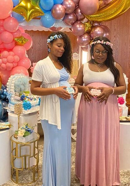 Two expectant mothers in blue and pink dresses and flower crowns in front of a balloon arch